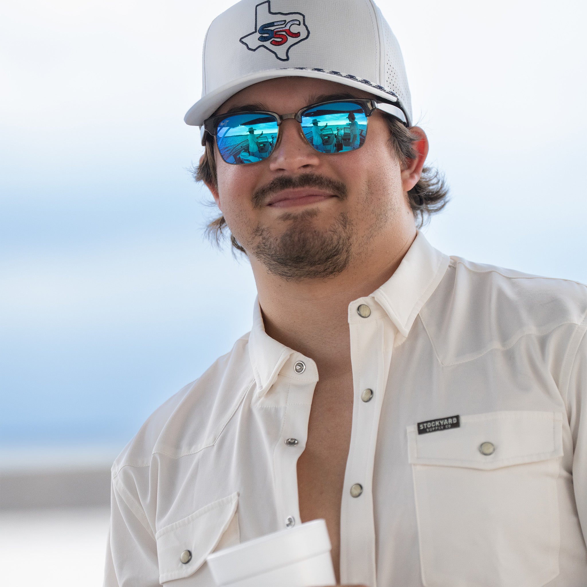 Man wearing a white cap with a logo and reflective sunglasses, standing against a clear sky.