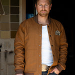 Eric Nelsen wearing "The Stockshow" Canvas Bomber jacket standing in a doorway with a cowboy hat in hand. 