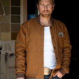 Eric Nelsen wearing "The Stockshow" Canvas Bomber jacket standing in a doorway with a cowboy hat in hand. 