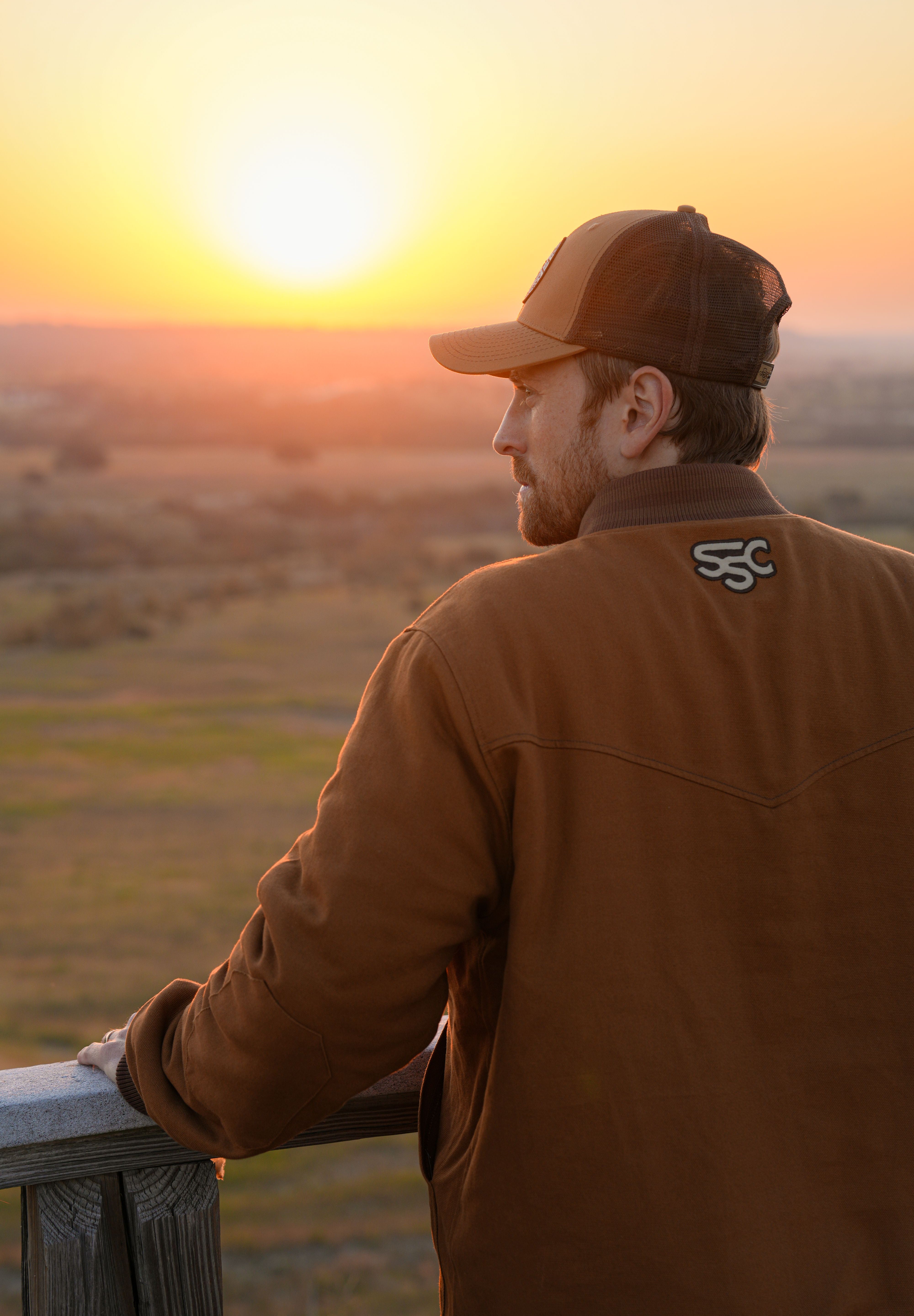 Eric Nelsen wearing "The Stockshow" bomber jacket, back towards the camera, looking out over a fence looking at the sunset. 