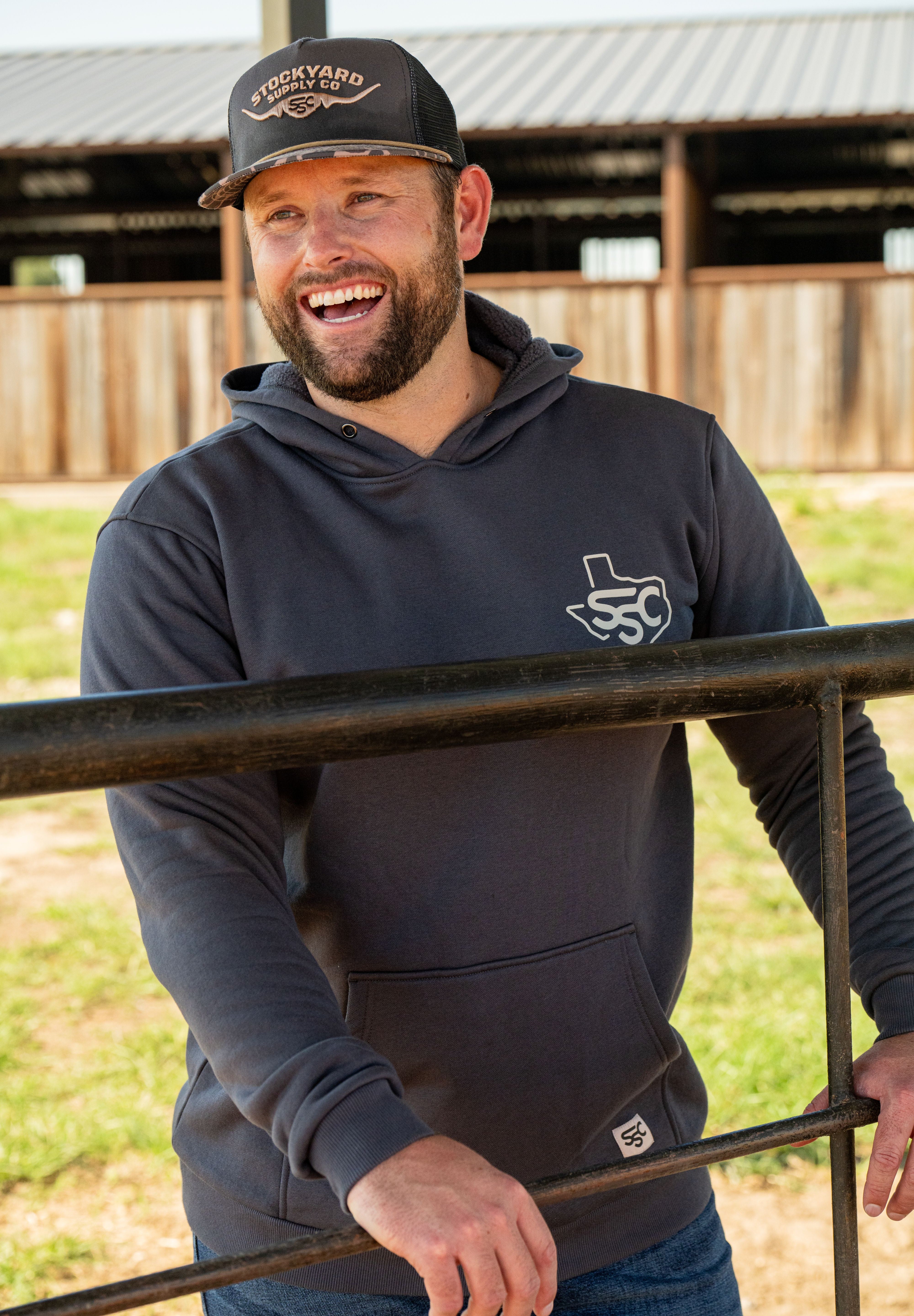 Ryan Dukes leaning on a fence with Stockyard Supply Co Charcoal hoodie. 