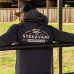 Ryan Dukes leaning on a fence, back towards the camera to show the back of his Stockyard Supply Co Charcoal hoodie. 