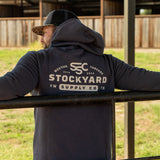 Ryan Dukes leaning on a fence, back towards the camera to show the back of his Stockyard Supply Co Charcoal hoodie. 