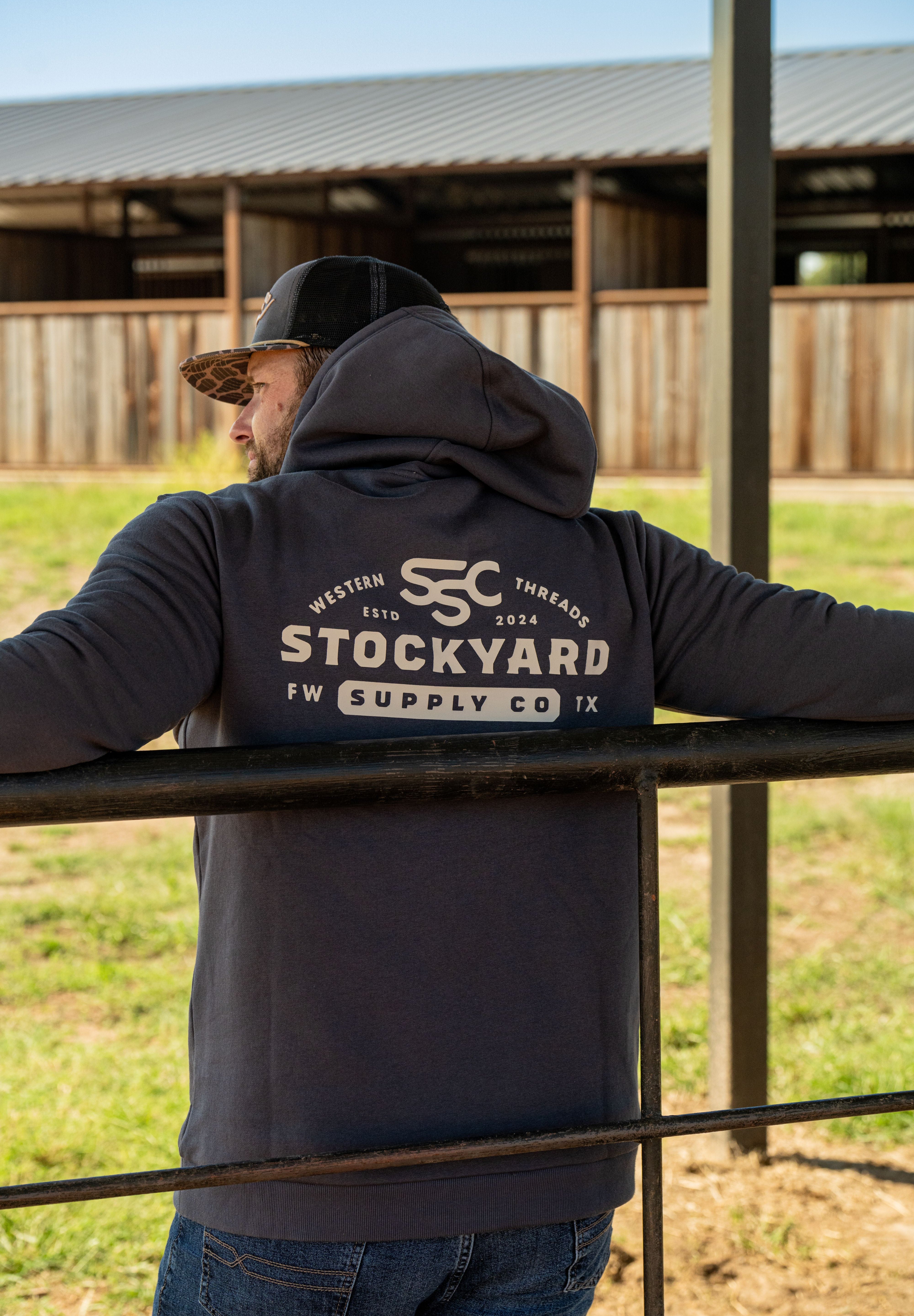 Ryan Dukes leaning on a fence, back towards the camera to show the back of his Stockyard Supply Co Charcoal hoodie. 