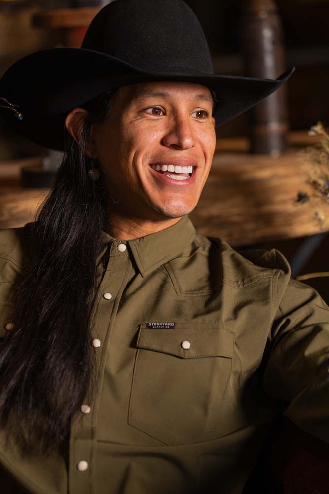 JP Guana sitting down wearing a black cowboy hat and the long sleeve olive green performance pearl snap from Stockyard Supply Co