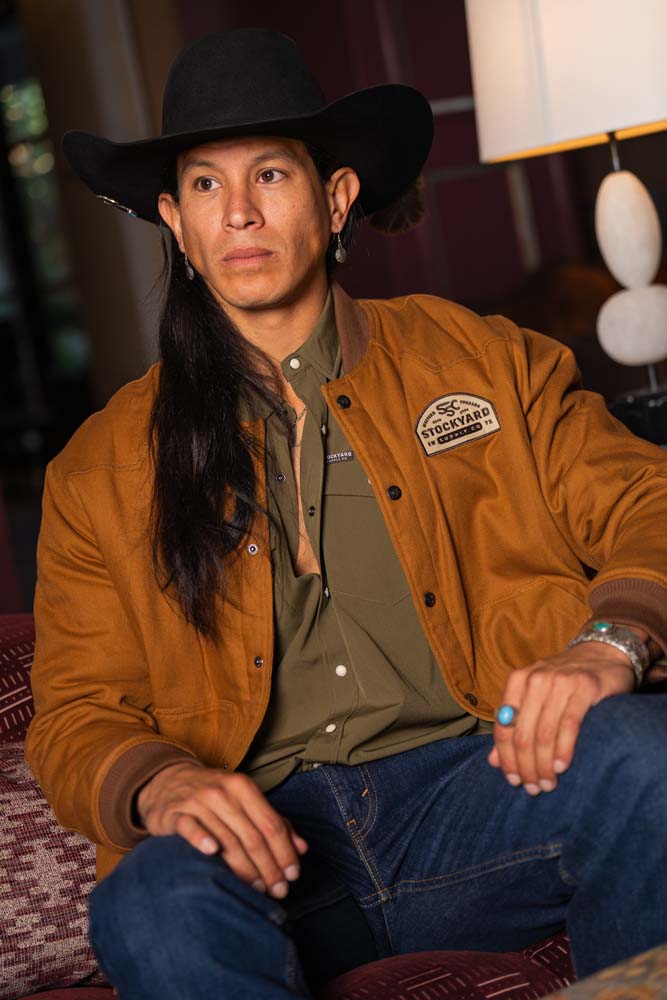 JP Guana sitting down wearing a black cowboy hat wearing a Stockyard Supply Co Olive pearl snap shirt under "The Stockshow" Canvas Bomber Jacket.