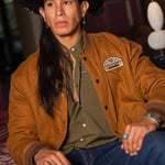 JP Guana sitting down wearing a black cowboy hat wearing a Stockyard Supply Co Olive pearl snap shirt under "The Stockshow" Canvas Bomber Jacket.