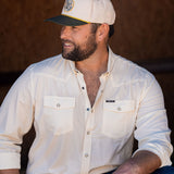 Ryan Dukes sitting on a bale of hay wearing blue jeans, the cream colored performance pearl snap, and The Duckster Stockyard Supply Co hat, cream colored panel hat with olive green bill. 