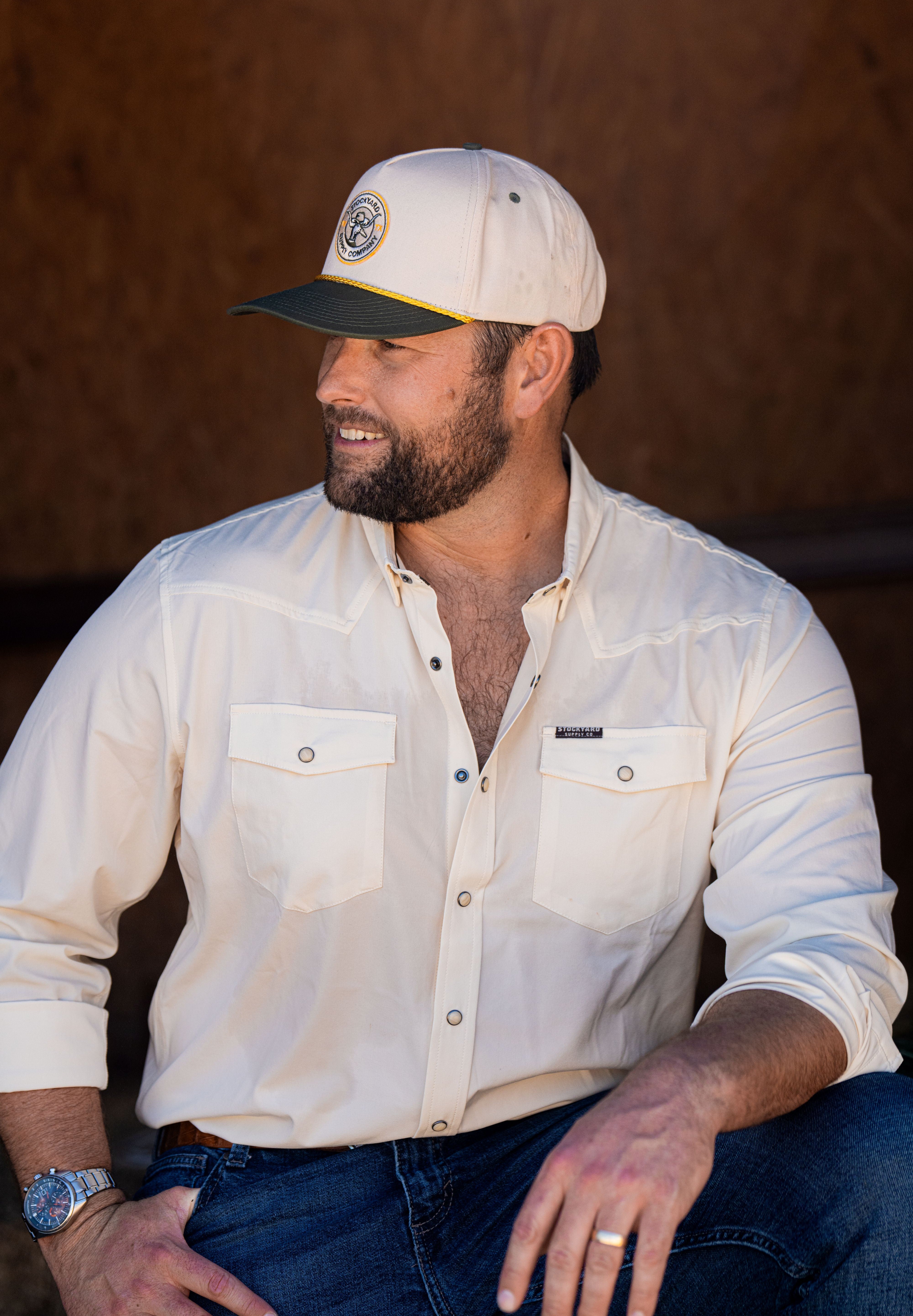 Ryan Dukes sitting on a bale of hay wearing blue jeans, the cream colored performance pearl snap, and The Duckster Stockyard Supply Co hat, cream colored panel hat with olive green bill. 