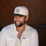 Ryan Dukes sitting on a bale of hay wearing blue jeans, the cream colored performance pearl snap, and The Duckster Stockyard Supply Co hat, cream colored panel hat with olive green bill. 