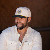 Ryan Dukes sitting on a bale of hay wearing blue jeans, the cream colored performance pearl snap, and The Duckster Stockyard Supply Co hat, cream colored panel hat with olive green bill. 