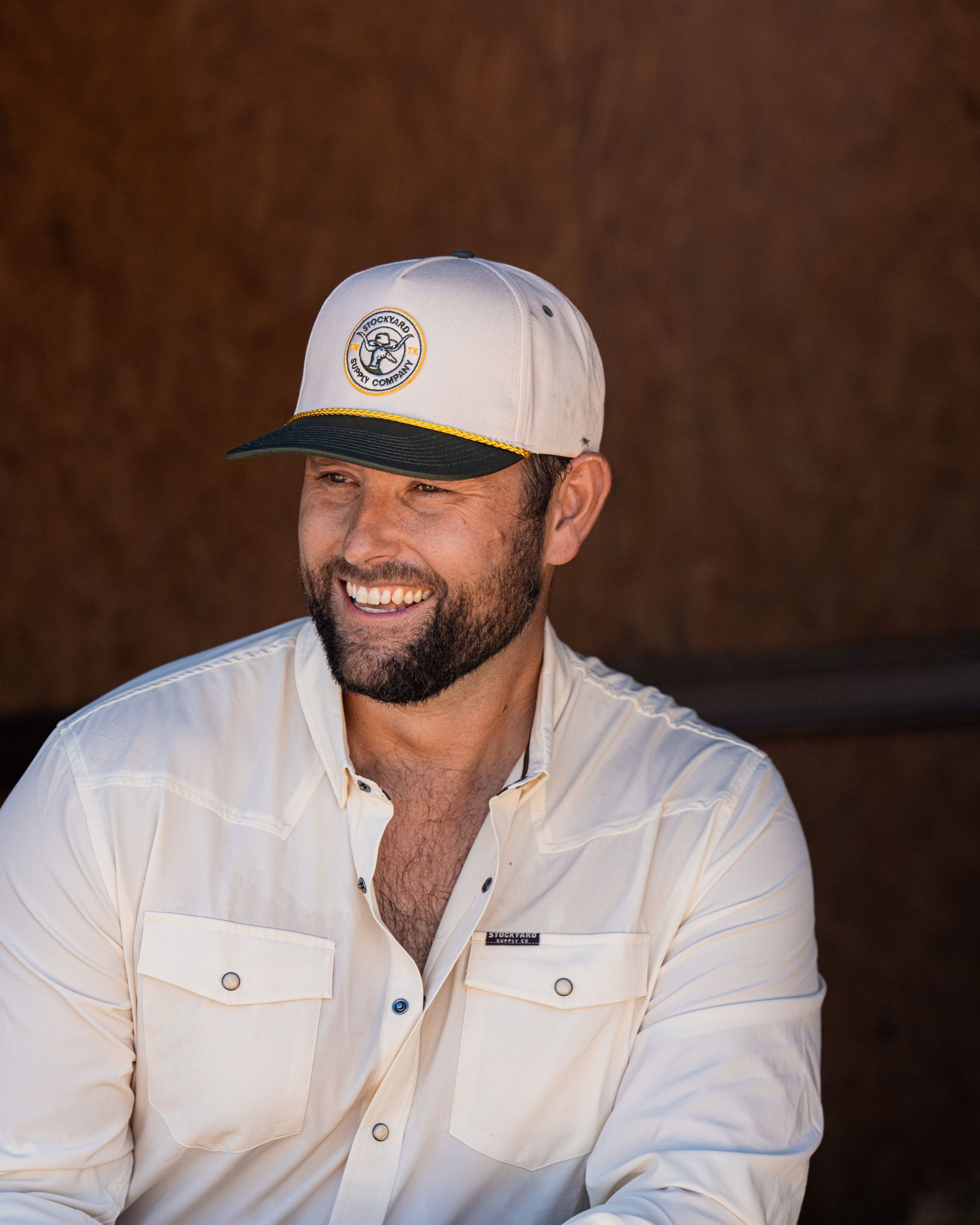 Ryan Dukes sitting on a bale of hay wearing blue jeans, the cream colored performance pearl snap, and The Duckster Stockyard Supply Co hat, cream colored panel hat with olive green bill. 