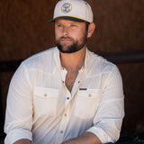 Ryan Dukes sitting on a bale of hay wearing blue jeans, the cream colored performance pearl snap, and The Duckster Stockyard Supply Co hat, cream colored panel hat with olive green bill. 