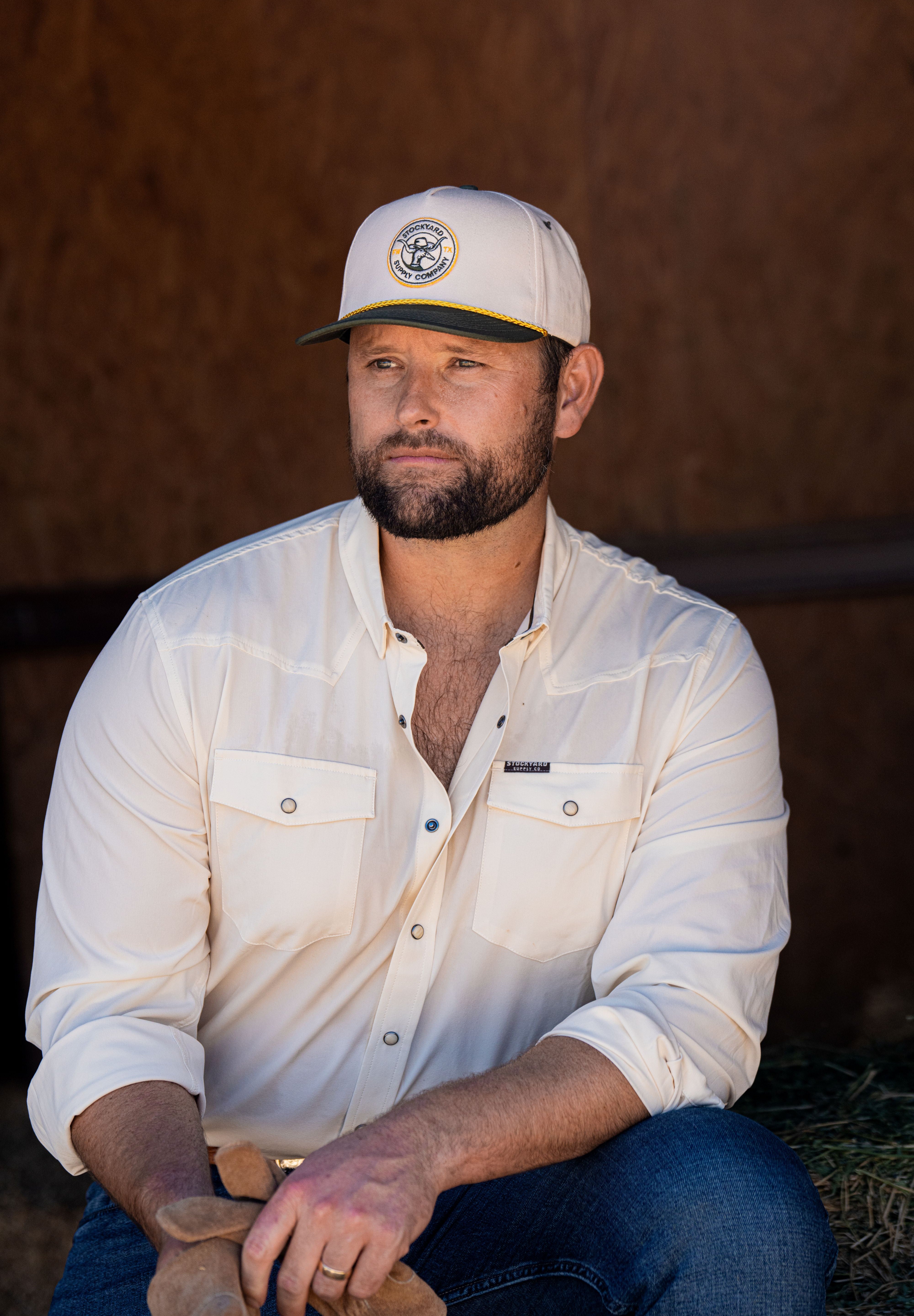 Ryan Dukes sitting on a bale of hay wearing blue jeans, the cream colored performance pearl snap, and The Duckster Stockyard Supply Co hat, cream colored panel hat with olive green bill. 