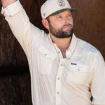 Ryan Dukes sitting on a bale of hay wearing blue jeans, the cream colored performance pearl snap, and The Duckster Stockyard Supply Co hat, cream colored panel hat with olive green bill. 