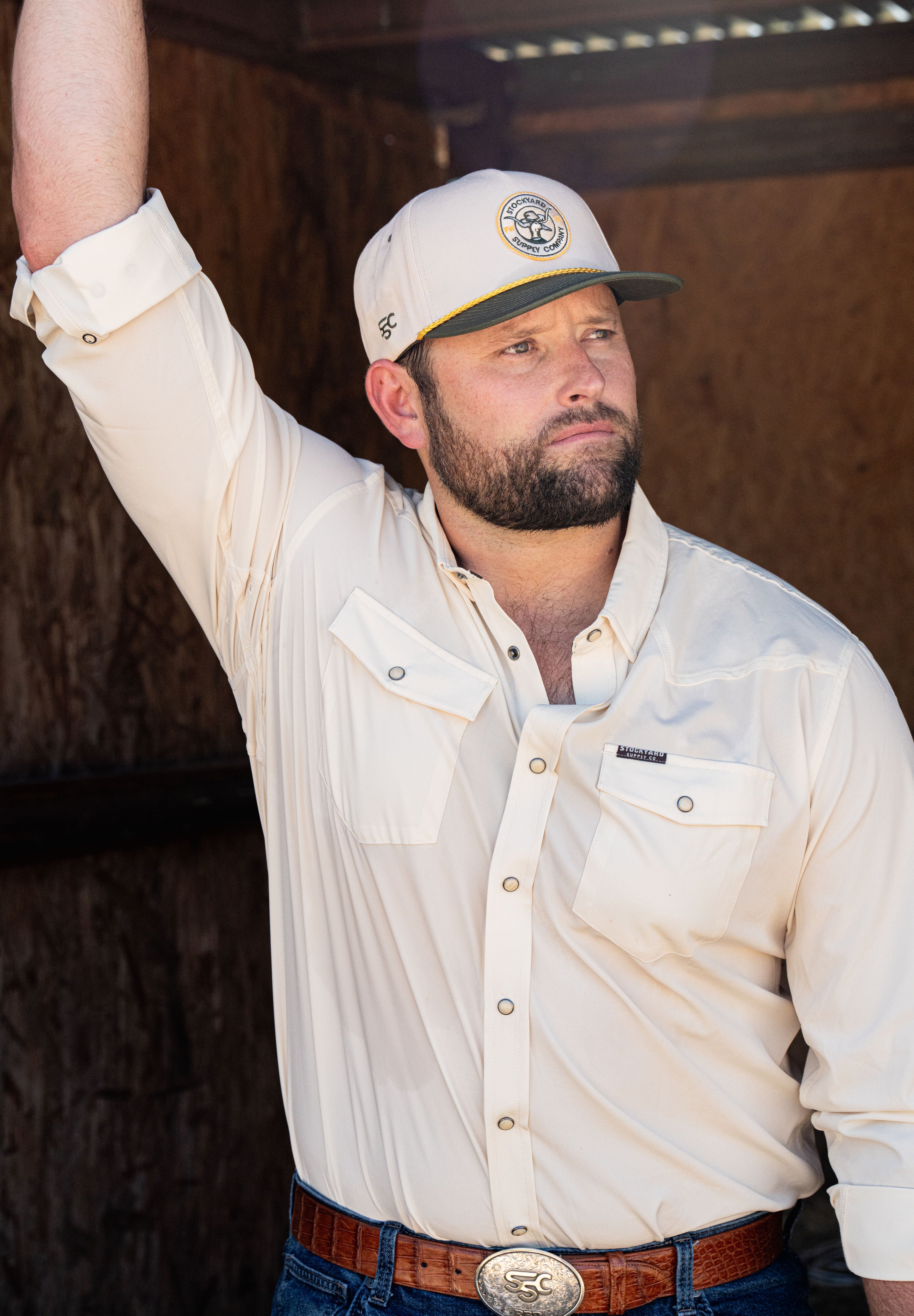 Ryan Dukes sitting on a bale of hay wearing blue jeans, the cream colored performance pearl snap, and The Duckster Stockyard Supply Co hat, cream colored panel hat with olive green bill. 