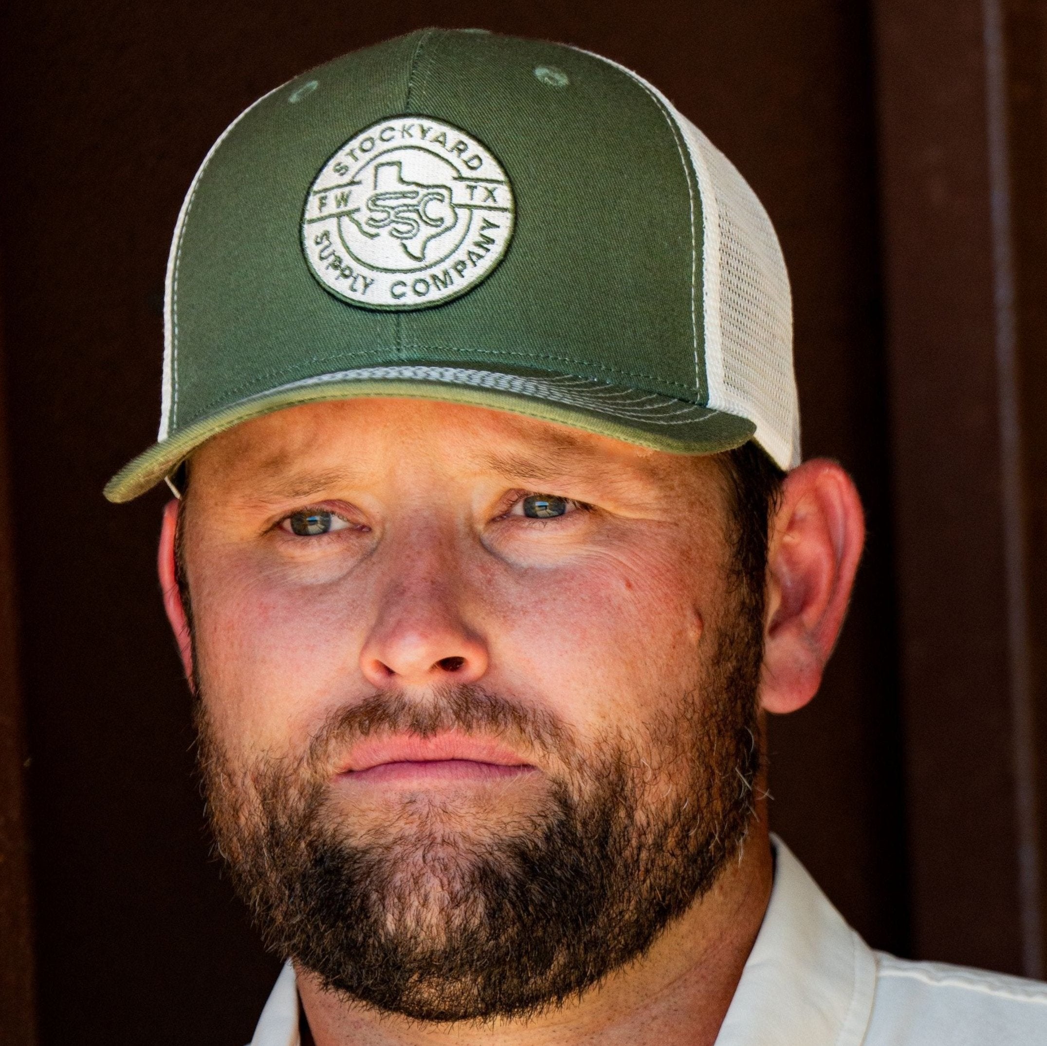 Ryan Dukes leaning against a barn door wearing the Everyday Jack Trucker Hat - Olive and Cream colored. The shirt he is wearing is the cream colored Stockyard Supply Co pearl snap performance button down.