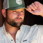 Ryan Dukes leaning against a barn door holding the brim of his hat - the Everyday Jack Trucker Hat, Olive and Cream colored. The shirt he is wearing is the cream colored Stockyard Supply Co pearl snap performance button down.