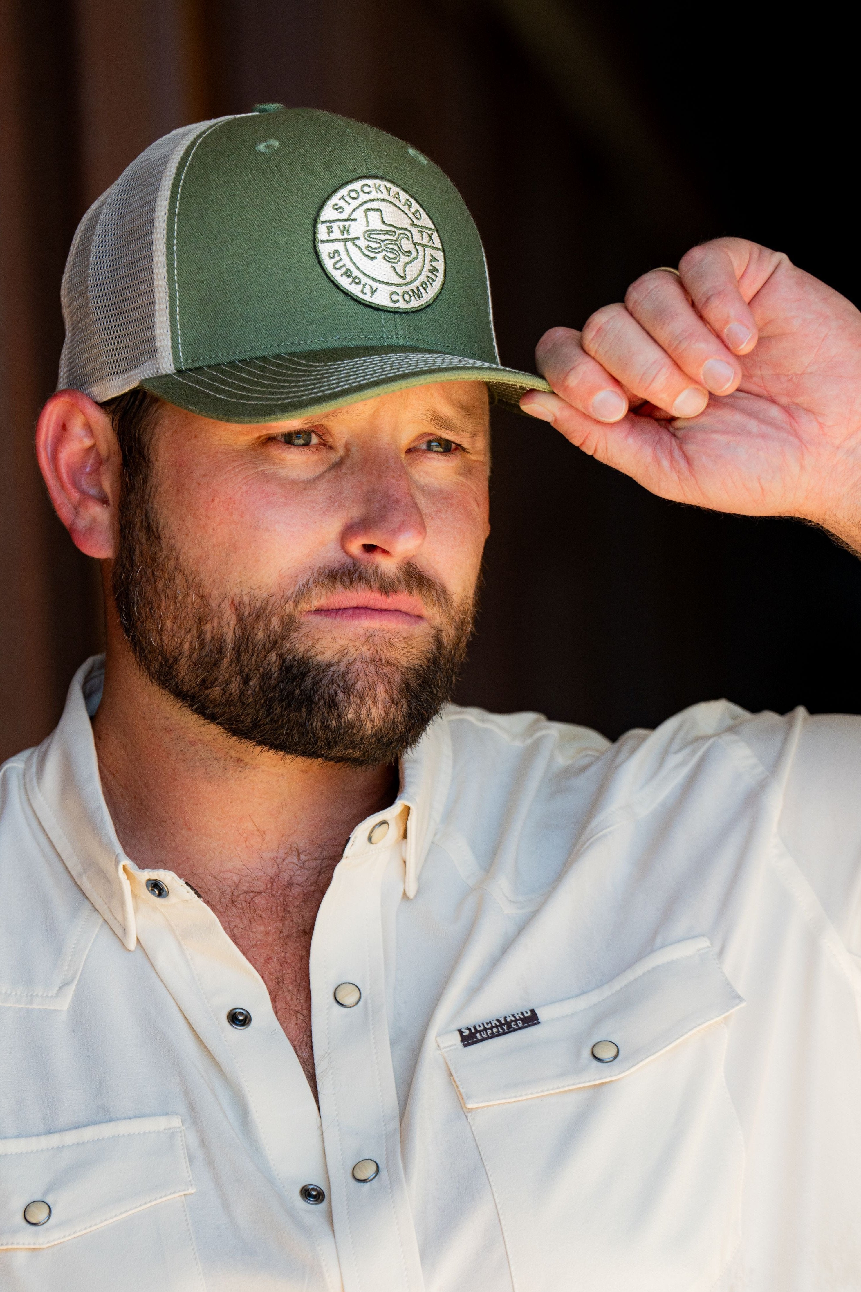 Ryan Dukes leaning against a barn door holding the brim of his hat - the Everyday Jack Trucker Hat, Olive and Cream colored. The shirt he is wearing is the cream colored Stockyard Supply Co pearl snap performance button down.