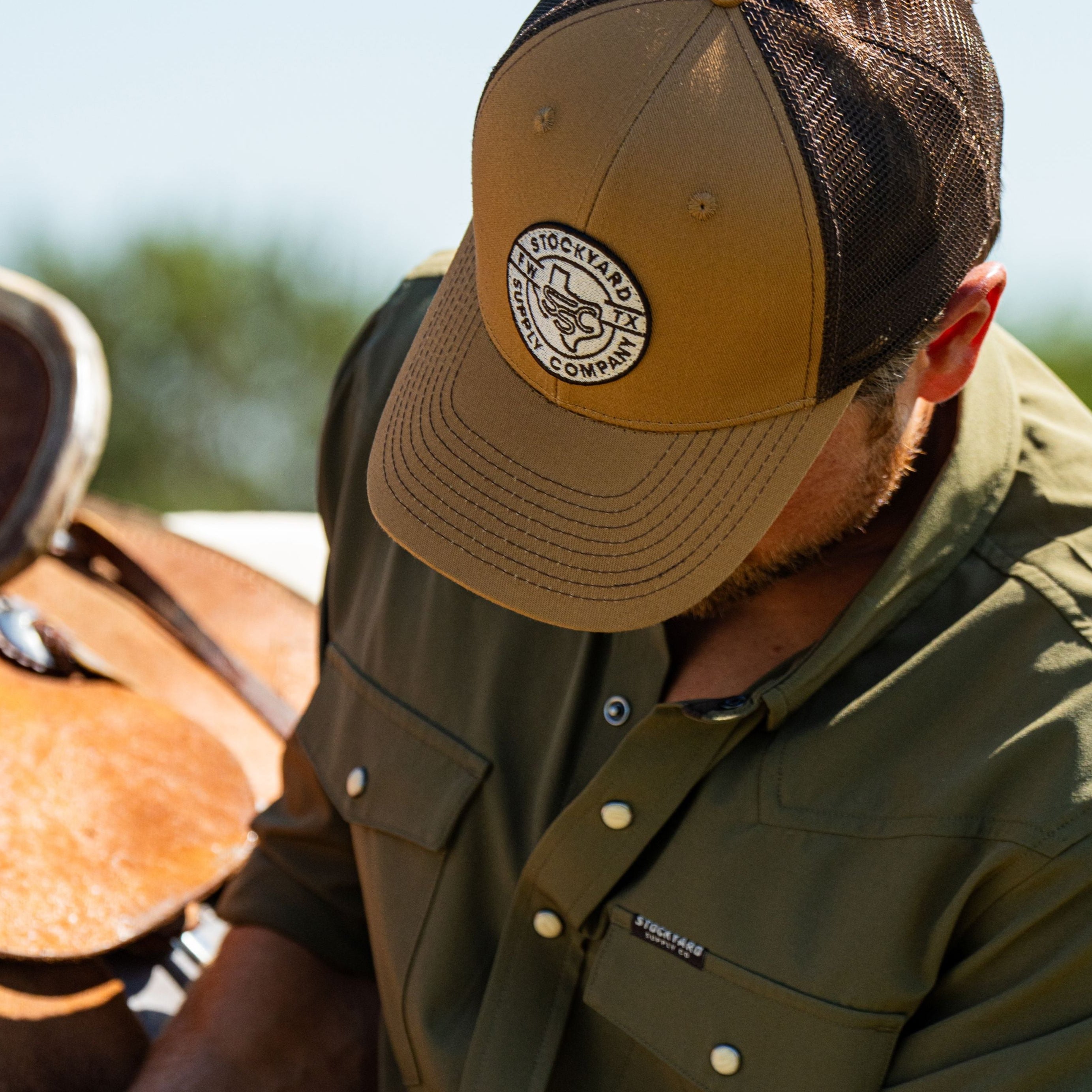 Eric Nelsen cinching down a saddle on a horse wearing the Everyday Jack Trucker Hat, Brown and Coyote colored. He is wearing blue jeans and the Stockyard Supply Co faded olive performance pearl snap button down.