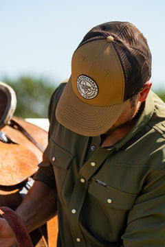 Eric Nelsen cinching down a saddle on a horse wearing the Everyday Jack Trucker Hat, Brown and Coyote colored. He is wearing blue jeans and the Stockyard Supply Co faded olive performance pearl snap button down.