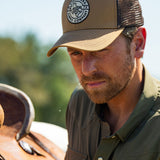 Eric Nelsen cinching down a saddle on a horse wearing the Everyday Jack Trucker Hat, Brown and Coyote colored. He is wearing blue jeans and the Stockyard Supply Co faded olive performance pearl snap button down.