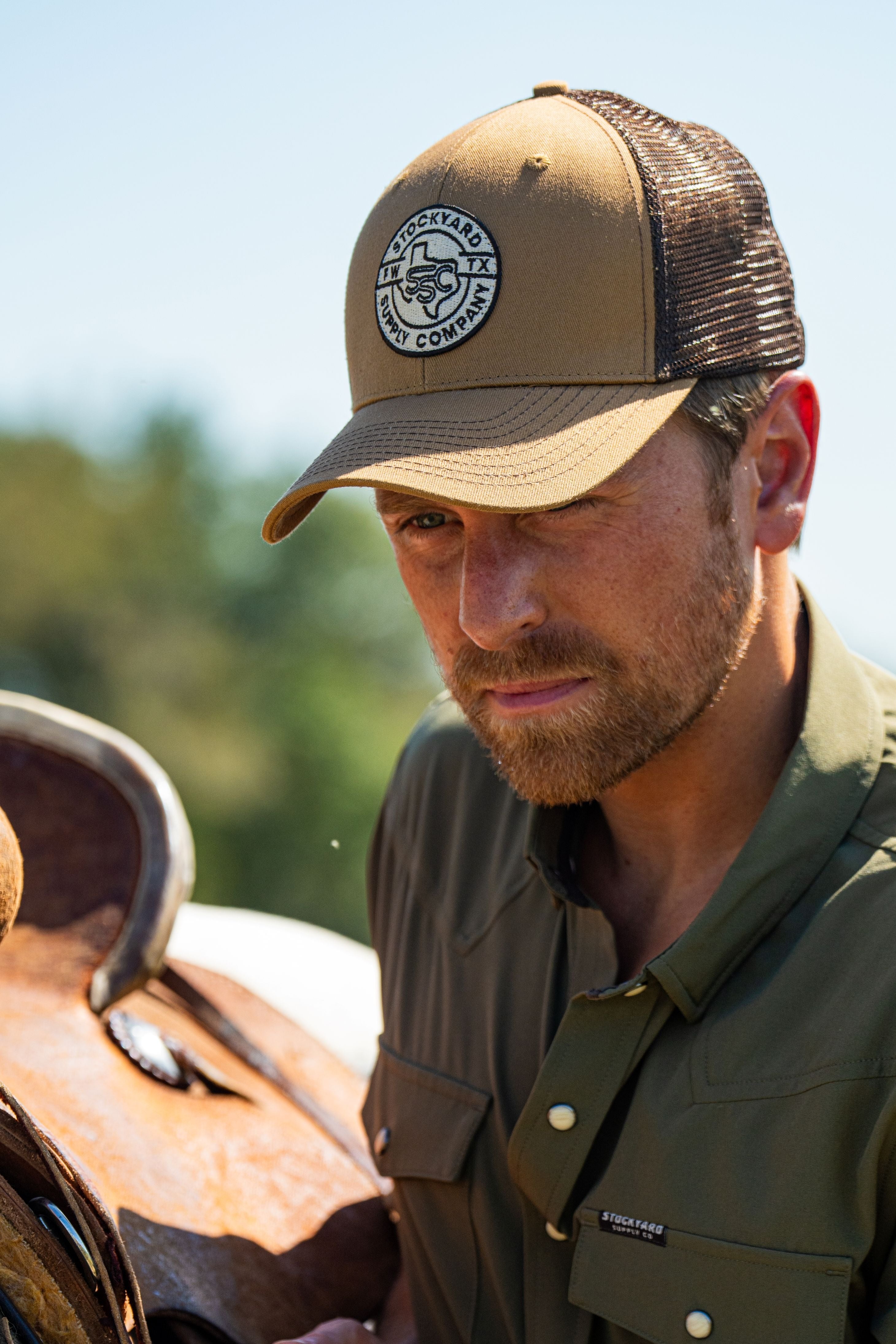 Eric Nelsen cinching down a saddle on a horse wearing the Everyday Jack Trucker Hat, Brown and Coyote colored. He is wearing blue jeans and the Stockyard Supply Co faded olive performance pearl snap button down.