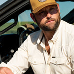 Ryan Dukes sitting in a wearing the Everyday Jack Trucker Hat, Brown and Coyote colored. He is wearing blue jeans and the Stockyard Supply Co cream performance pearl snap button down.