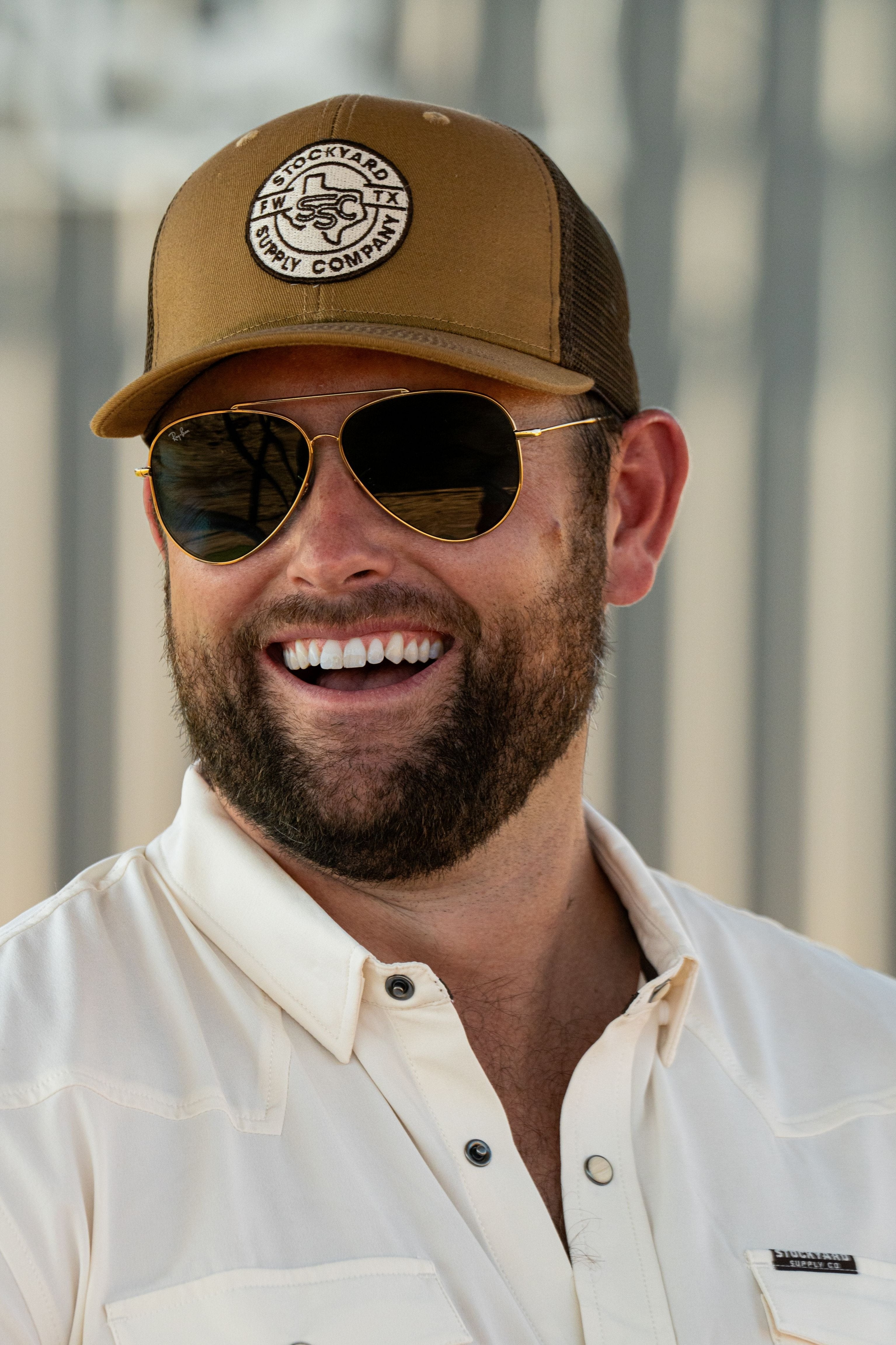 Ryan Dukes laughing wearing the Everyday Jack Trucker Hat, Brown and Coyote colored with aviator sunglasses. He is wearing the Stockyard Supply Co cream performance pearl snap button down.