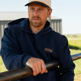 Eric Nelsen wearing a cream and navy Stockyard Supply Co hat with the Navy Performance Western Hoodie leaning on a metal fence. 