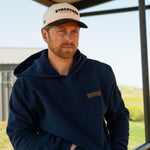 Eric Nelsen wearing a cream and navy Stockyard Supply Co hat with the Navy Performance Western Hoodie sitting on top of a metal fence with his hands in the pockets.