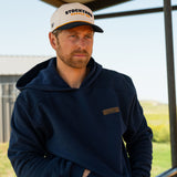 Eric Nelsen wearing a cream and navy Stockyard Supply Co hat with the Navy Performance Western Hoodie sitting on top of a metal fence with his hands in the pockets.