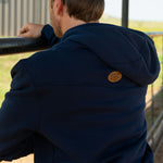 Eric Nelsen wearing a cream and navy Stockyard Supply Co hat with the Navy Performance Western Hoodie leaning on a metal fence facing away from the camera to show the back of the hoodie. 