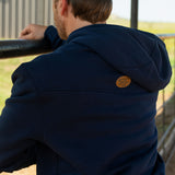 Eric Nelsen wearing a cream and navy Stockyard Supply Co hat with the Navy Performance Western Hoodie leaning on a metal fence facing away from the camera to show the back of the hoodie. 