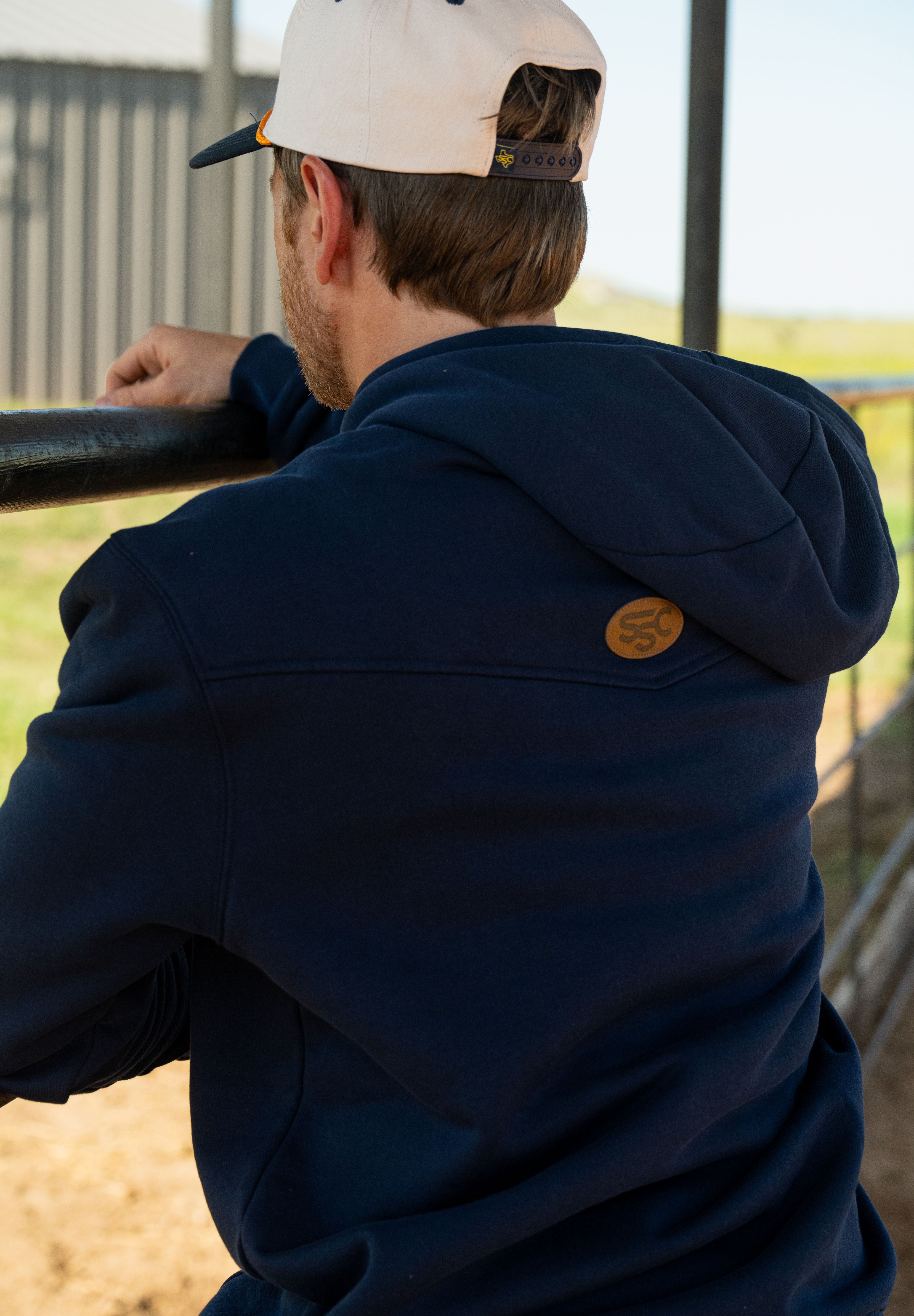 Eric Nelsen wearing a cream and navy Stockyard Supply Co hat with the Navy Performance Western Hoodie leaning on a metal fence facing away from the camera to show the back of the hoodie. 