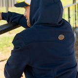 Eric Nelsen wearing a cream and navy Stockyard Supply Co hat with the Navy Performance Western Hoodie leaning on a metal fence with the hoodie on top of his head facing away from the camera. 