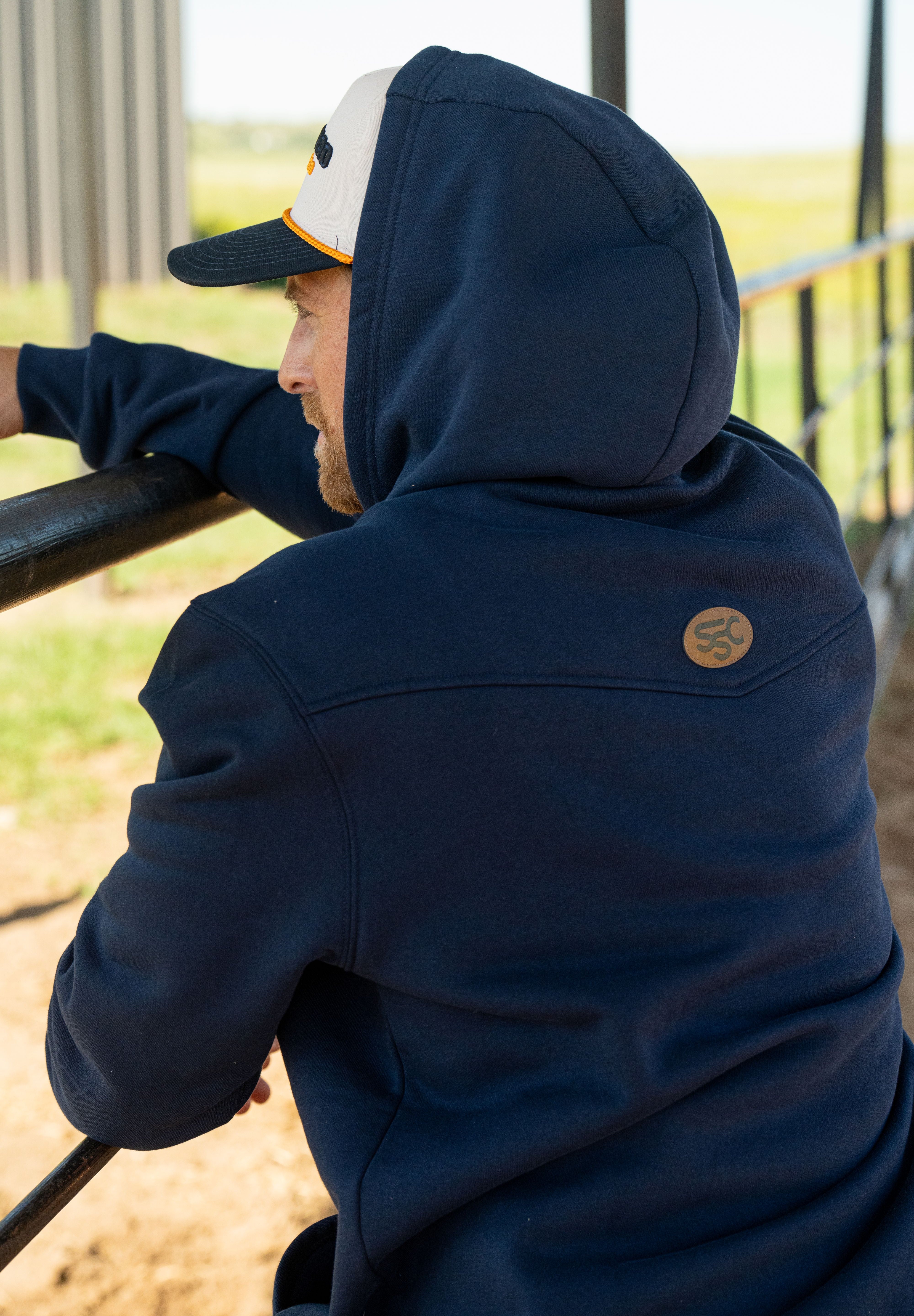 Eric Nelsen wearing a cream and navy Stockyard Supply Co hat with the Navy Performance Western Hoodie leaning on a metal fence with the hoodie on top of his head facing away from the camera. 