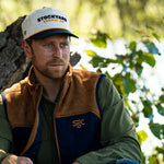 Eric Nelsen leaning against a Tree with a white and navy Stockyard Supply Co hat wearing a Stockyard Supply Co Rodeo King Vest - Navy and Coyote colors with the faded olive pearl snap performance shirt.