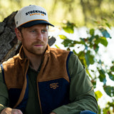 Eric Nelsen leaning against a Tree with a white and navy Stockyard Supply Co hat wearing a Stockyard Supply Co Rodeo King Vest - Navy and Coyote colors with the faded olive pearl snap performance shirt.