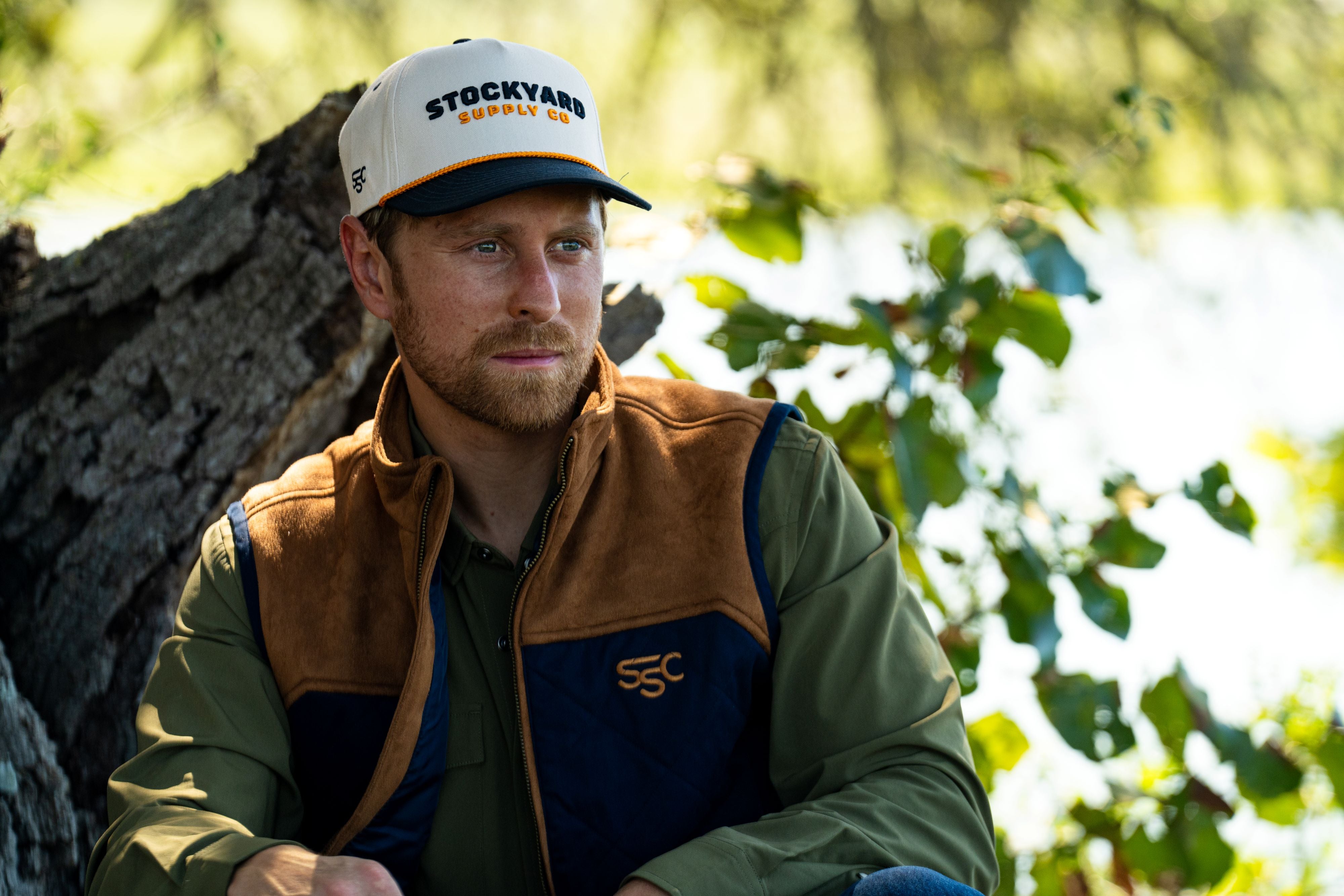 Eric Nelsen leaning against a Tree with a white and navy Stockyard Supply Co hat wearing a Stockyard Supply Co Rodeo King Vest - Navy and Coyote colors with the faded olive pearl snap performance shirt.