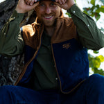 Eric Nelsen sitting against a Tree with a white and navy Stockyard Supply Co holding the brim of the hat wearing a Stockyard Supply Co Rodeo King Vest - Navy and Coyote colors with the faded olive pearl snap performance shirt.