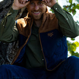 Eric Nelsen sitting against a Tree with a white and navy Stockyard Supply Co holding the brim of the hat wearing a Stockyard Supply Co Rodeo King Vest - Navy and Coyote colors with the faded olive pearl snap performance shirt.