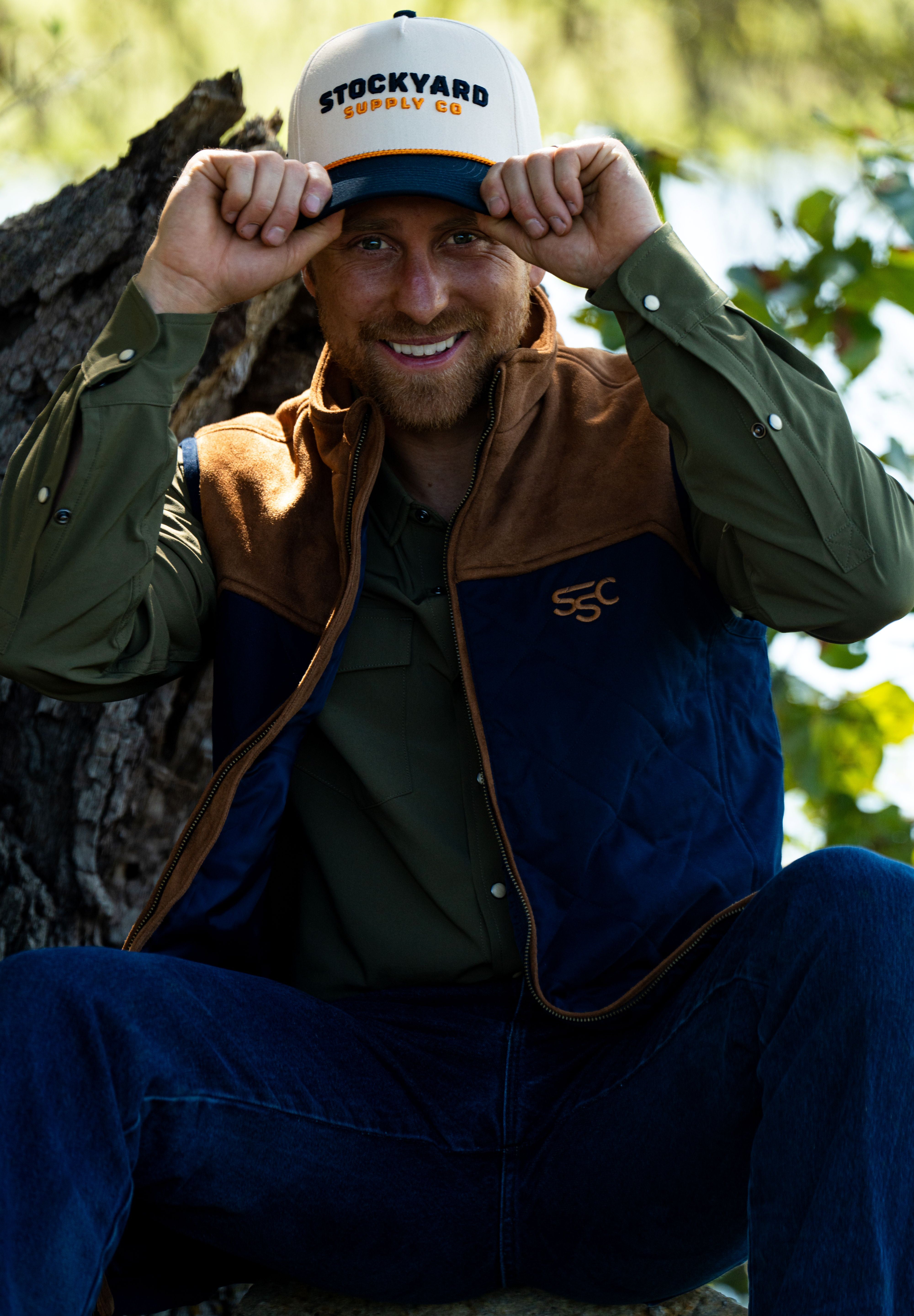 Eric Nelsen sitting against a Tree with a white and navy Stockyard Supply Co holding the brim of the hat wearing a Stockyard Supply Co Rodeo King Vest - Navy and Coyote colors with the faded olive pearl snap performance shirt.