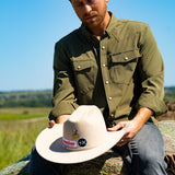 Eric Nelsen wearing a faded olive pearl snap button down shirt sitting on a hay bale holding a cowboy hat