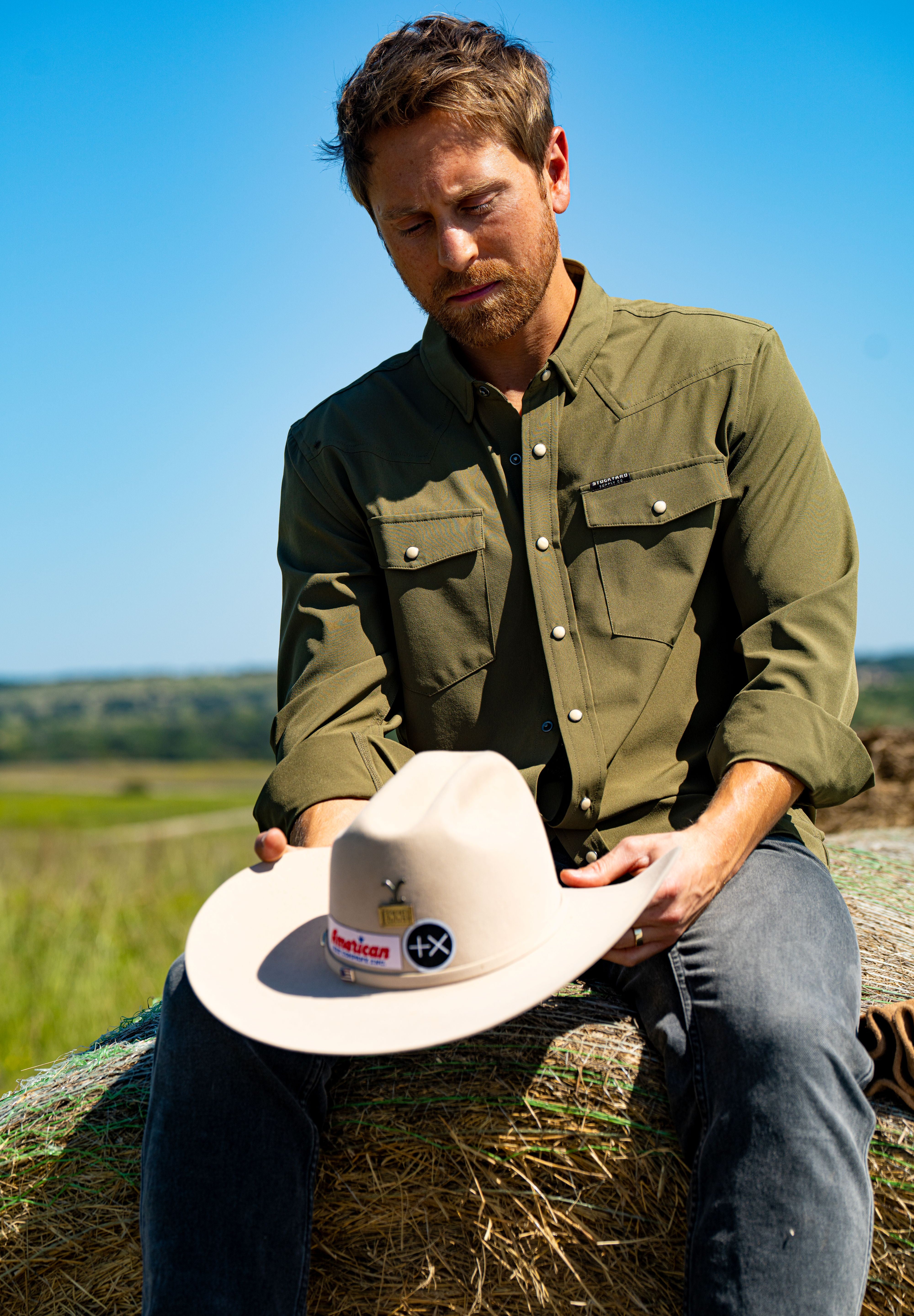 Eric Nelsen wearing a faded olive pearl snap button down shirt sitting on a hay bale holding a cowboy hat