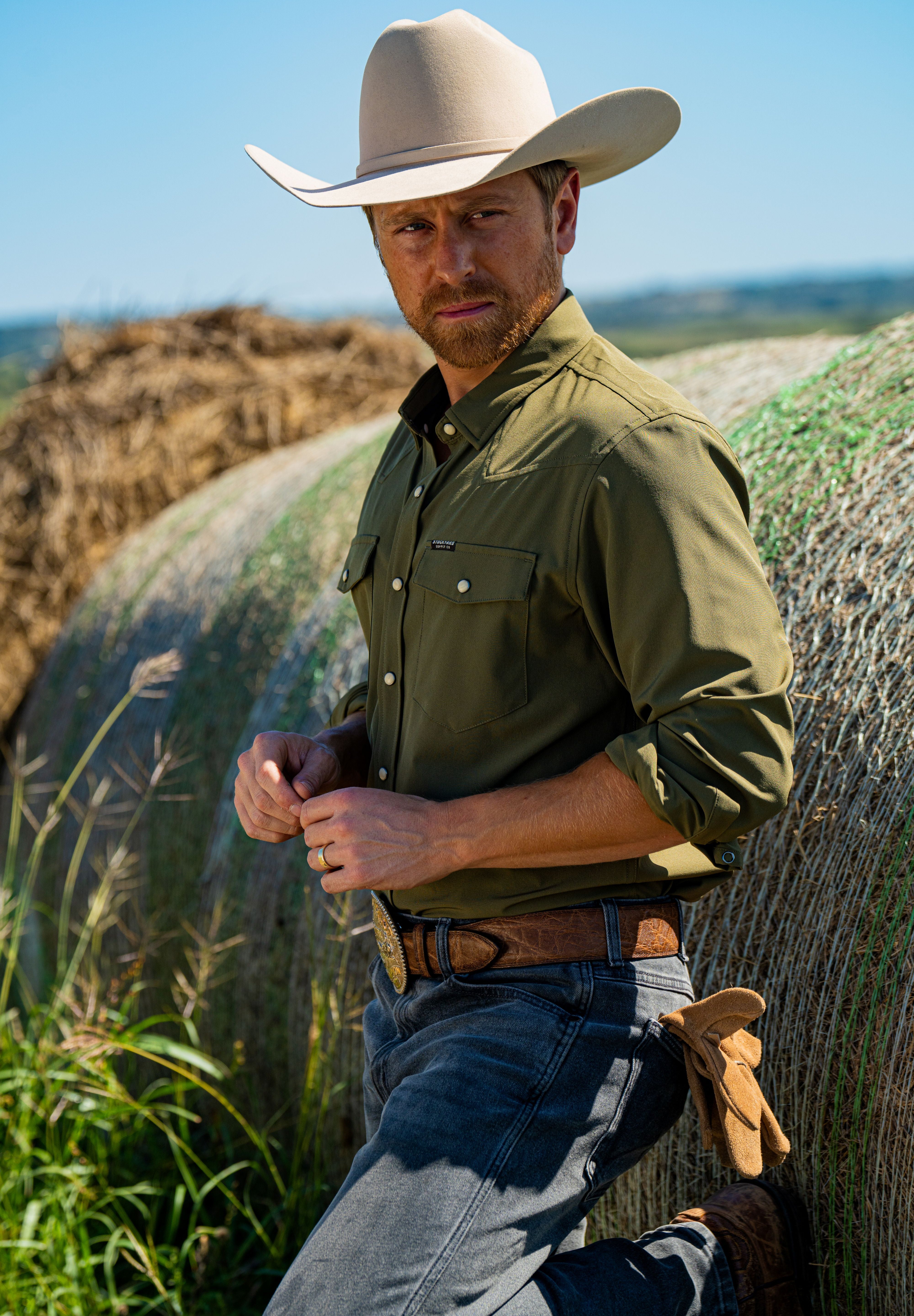 Eric Nelsen wearing a faded olive pearl snap button down shirt leaning against a hay bale