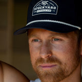 Eric Nelsen wearing The Champion Suede leather navy hat in a white tank top leaning up against a door.