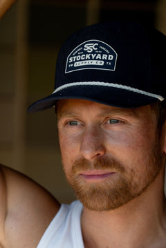 Eric Nelsen wearing The Champion Suede leather navy hat in a white tank top leaning up against a door.