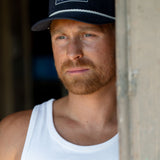 Eric Nelsen wearing The Champion Navy Suede leather hat in a white tank top leaning up against a door.