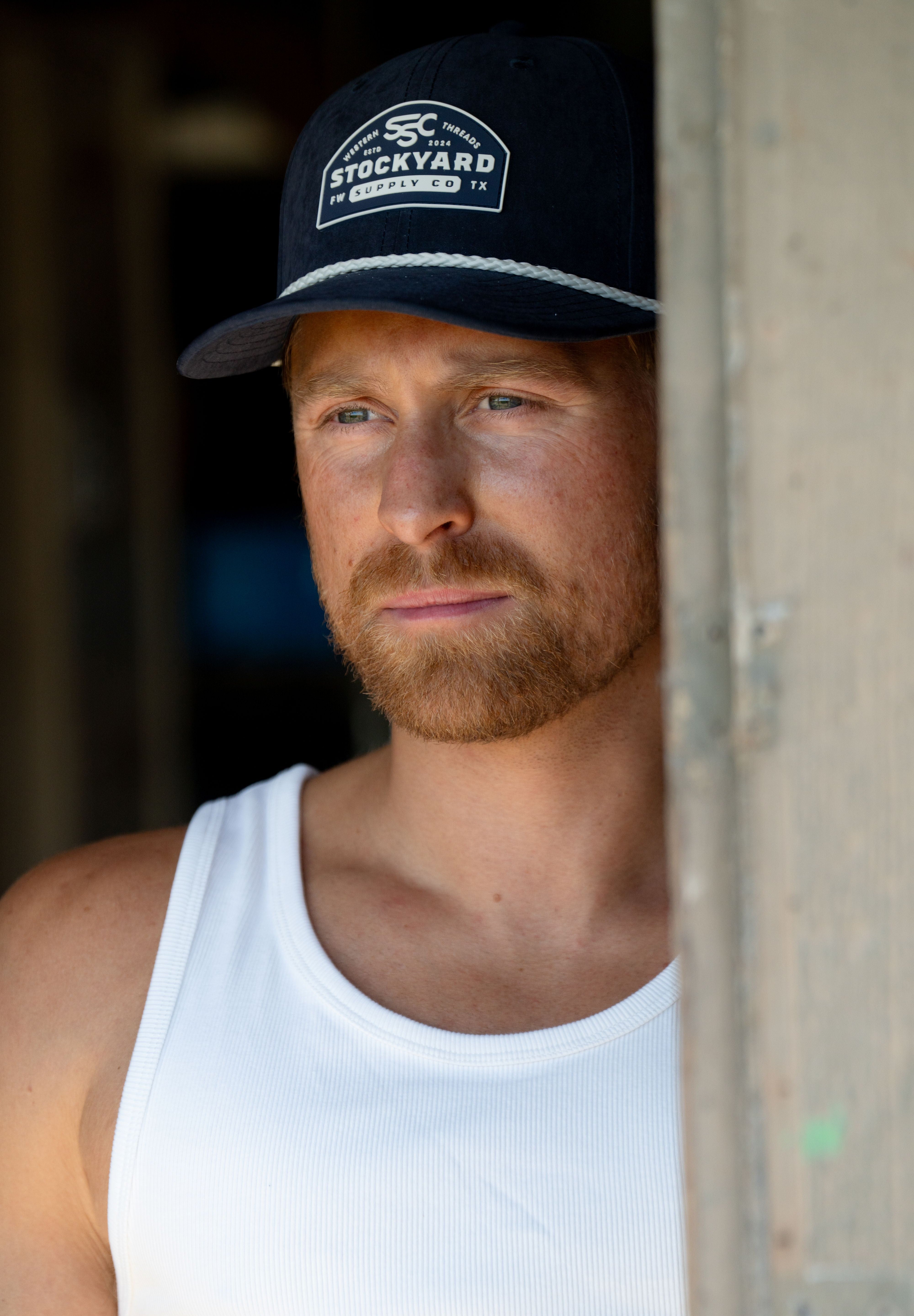 Eric Nelsen wearing The Champion Navy Suede leather hat in a white tank top leaning up against a door.